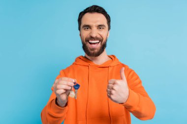 cheerful bearded man in orange hoodie holding key and showing thumb up isolated on blue