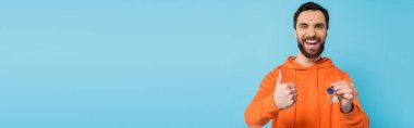 excited brunette man in orange hoodie showing like gesture while holding key isolated on blue, banner
