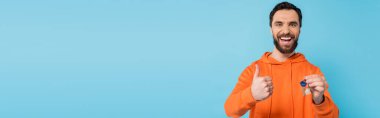 excited brunette man in orange hoodie showing like gesture while holding key isolated on blue, banner clipart
