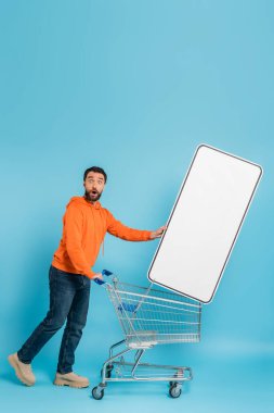 full length of amazed man looking at camera near shopping cart with huge phone template on blue background
