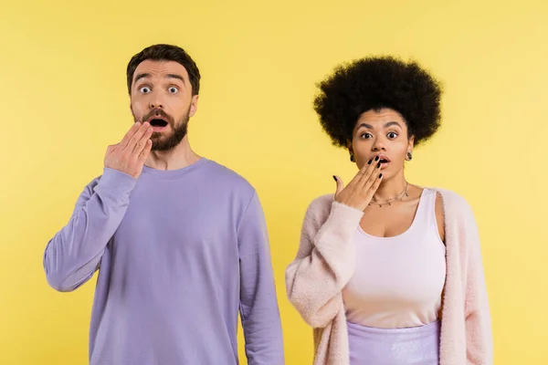 thrilled interracial couple covering open mouth with hands and looking at camera isolated on yellow