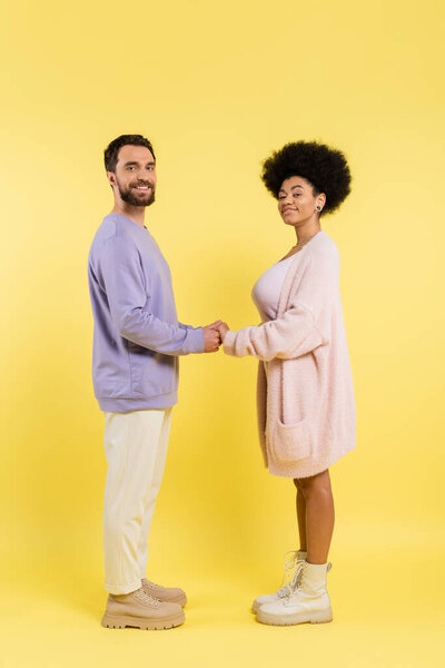 full length of interracial couple in stylish clothes holding hands and looking at camera on yellow background