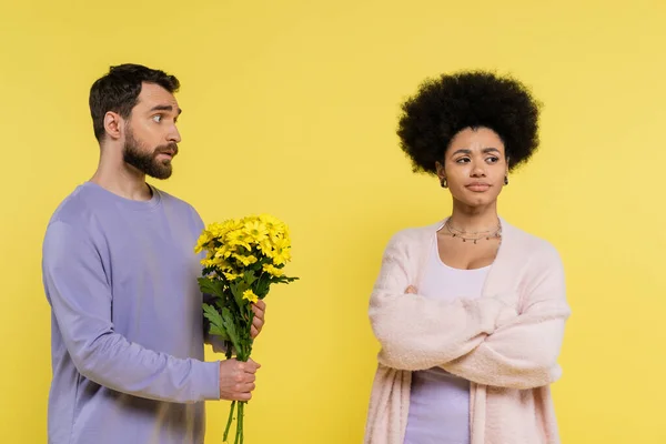 discouraged man holding flowers near offended african american woman standing with crossed arms isolated on yellow