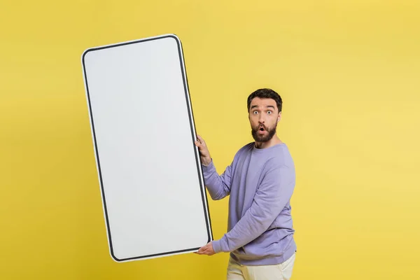 stock image thrilled bearded man holding white template of mobile phone and looking at camera isolated on yellow