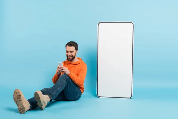 stock image full length of smiling bearded man messaging on smartphone while sitting near huge phone template on blue background