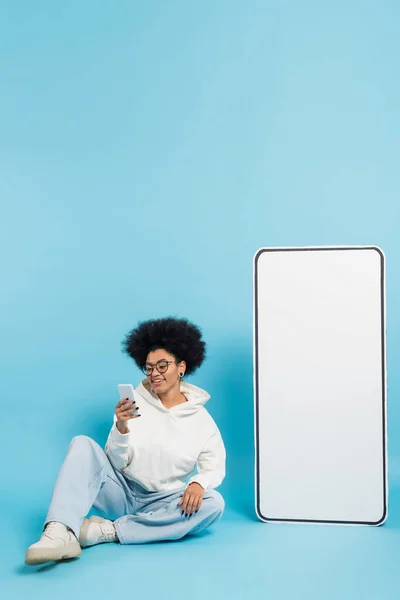 stock image full length of happy african american woman in eyeglasses sitting with smartphone near huge phone template on blue background