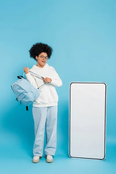 stock image full length of smiling african american student with backpack looking at camera near empty phone template on blue background