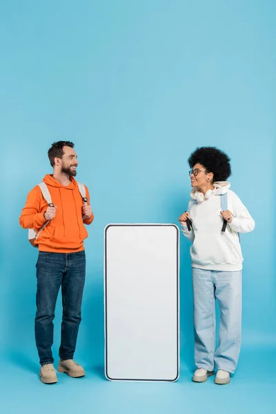 stock image cheerful multiethnic students with backpacks looking at each other near mobile phone mock-up on blue background