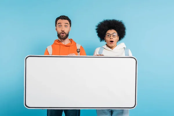 stock image amazed interracial students looking at camera near white phone template isolated on blue