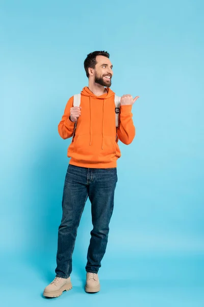 stock image full length of joyful student with backpack looking away and pointing with thumb on blue background