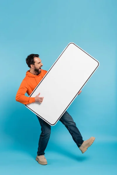 stock image full length of excited man holding huge template of smartphone on blue background