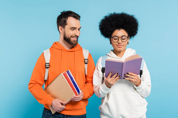 кудрявая африканская американка в очках читает книгу рядом с бородатым студентом с блокнотами, изолированными на голубом