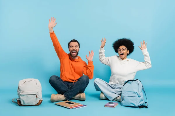 excited interracial students screaming and waving hands while sitting near backpacks and notebooks on blue background