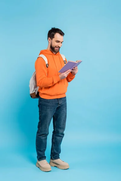 stock image full length of smiling bearded student in orange hoodie and jeans reading book on blue 