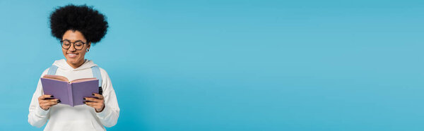  счастливая африканская американская студентка в модной толстовке и очках, читающая книгу, изолированную на синем, баннере