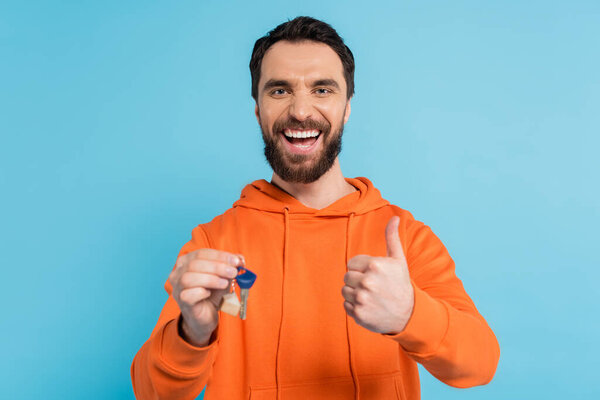 весёлый бородатый мужчина в оранжевой толстовке держит ключ и показывает большой палец на голубом