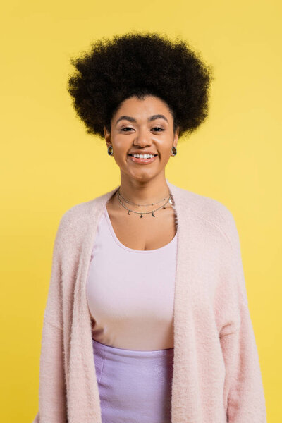cheerful african american woman in soft cardigan and silver necklaces looking at camera isolated on yellow