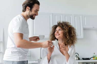 happy young man giving cup of coffee to cheerful girlfriend in kitchen 