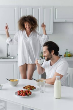 cheerful man with closed eyes drinking coffee near girlfriend dancing on blurred background 
