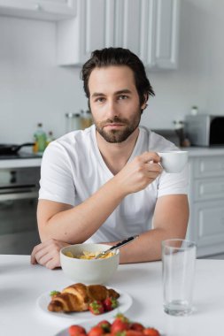 bearded man holding cup of coffee near tasty breakfast on table in kitchen 