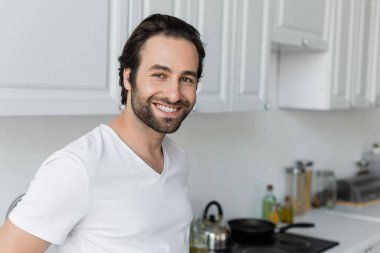 happy and bearded man in white t-shirt looking at camera in kitchen 