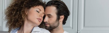 bearded man seducing sexy and curly woman with closed eyes in kitchen, banner 