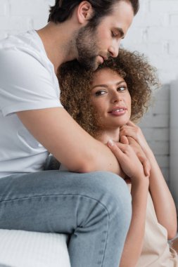 bearded man in jeans and white t-shirt hugging curly woman in bedroom 