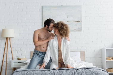 shirtless man in jeans looking at cheerful and curly woman in bedroom 