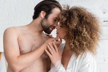 shirtless man touching face and kissing curly woman in white satin robe 
