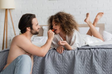 shirtless man feeding sensual woman in silk robe with ripe strawberry 