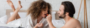 shirtless man feeding sensual woman in silk robe with ripe strawberry, banner 