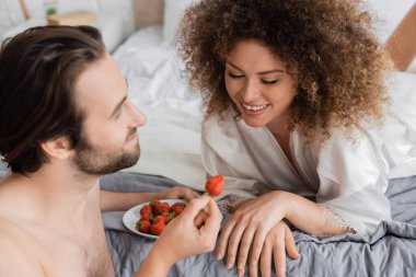 shirtless man holding ripe strawberry near cheerful young woman in silk robe