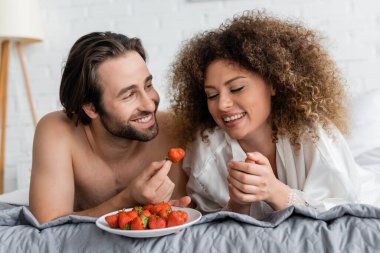 cheerful and young man holding fresh strawberry near happy girlfriend on bed 