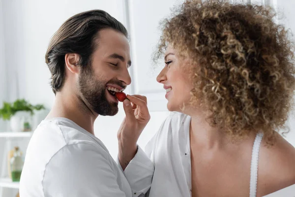 curly woman feeding cheerful boyfriend with fresh strawberry