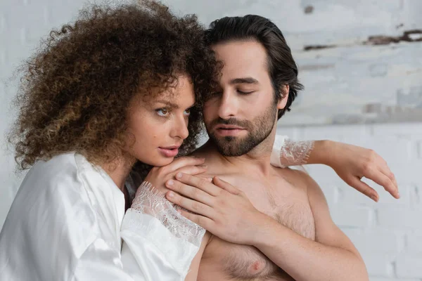 curly woman in silk robe hugging shirtless boyfriend at home