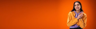 Smiling woman in hoodie and jacket looking at camera isolated on orange, banner 