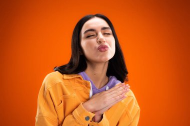 Young brunette woman pouting lips and closing eyes isolated on orange