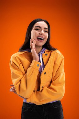 Excited brunette woman in jacket and hoodie looking at camera isolated on orange