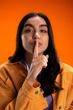 Portrait of brunette woman showing secret gesture isolated on orange