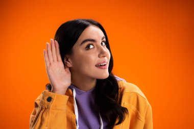 Young woman in jacket listening while holding hand near ear isolated on orange