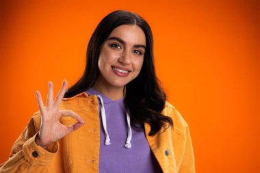 Portrait of smiling brunette woman showing ok sign isolated on orange