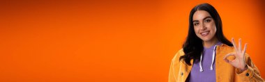 Young woman smiling and showing okay gesture at camera isolated on orange, banner 