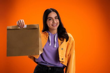 Cheerful woman holding paper bag isolated on orange