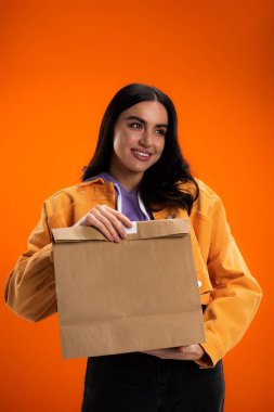 Smiling woman in casual clothes holding paper bag isolated on orange