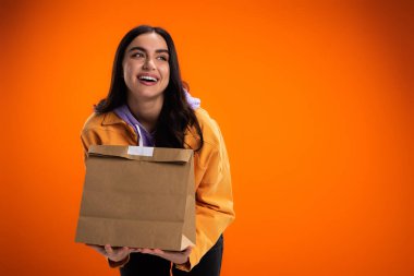 Cheerful woman holding paper bag with food isolated on orange