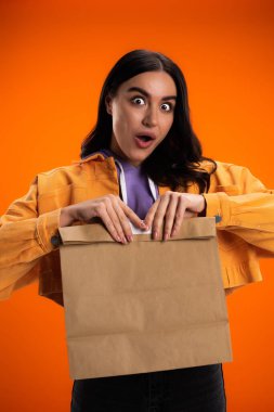 Shocked brunette woman holding paper bag isolated on orange