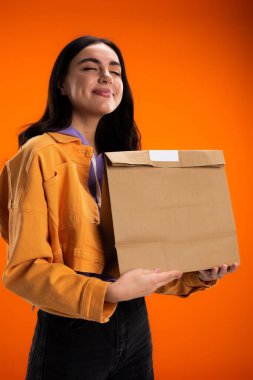 Joyful young woman with closed eyes holding paper bag isolated on orange