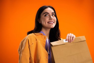 Dreamy young woman in jacket holding paper bag isolated on orange