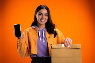 Smiling woman holding paper bag and smartphone with blank screen isolated on orange