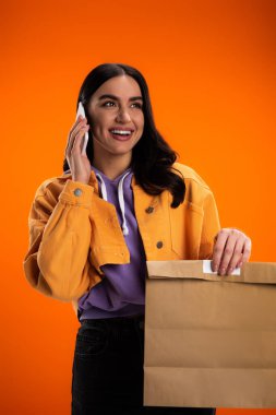 Cheerful brunette woman talking on smartphone and holding paper bag isolated on orange